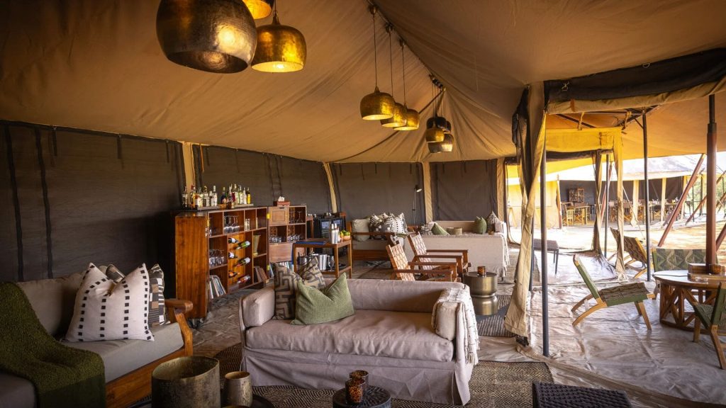 Olakira Migration Camp: Lounge tent interior: seating, wooden bar shelves, lantern lights, canvas walls opening to the outdoor dining area
