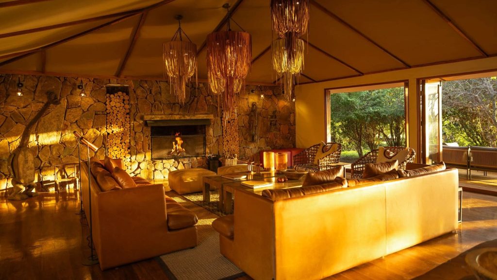 Sayari Camp: Main lounge with leather sofas, stone fireplace, woven pendant lights and open doors to the woodland